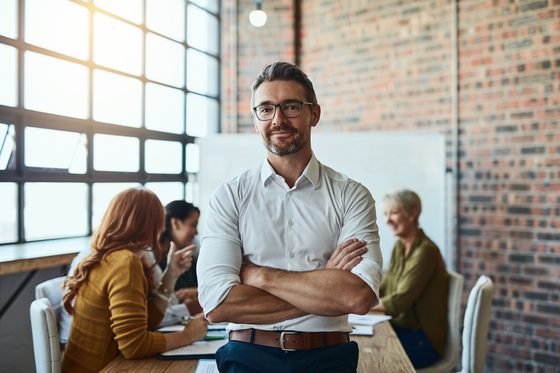 Un professionnel souriant, les bras croisés, dans un bureau, tandis que ses collègues collaborent en arrière-plan.