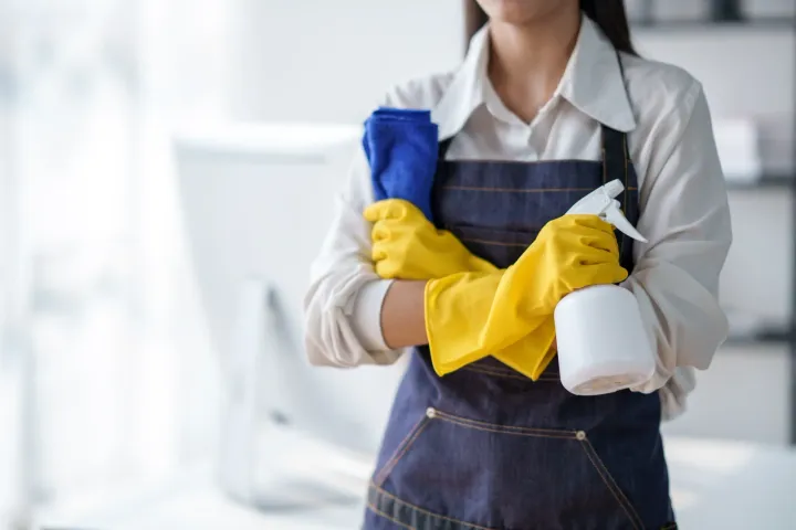 Eine Frau in einer blauen Schürze und gelben Handschuhen hält eine Sprühflasche und ein Tuch.