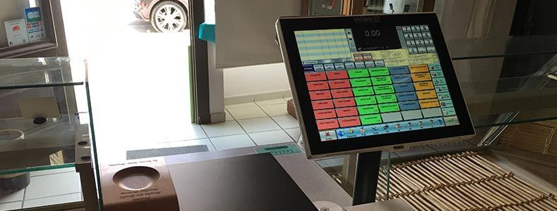 Un terminal de point de vente à écran tactile est posé sur un comptoir dans un magasin, avec vue sur une voiture garée à l'extérieur.