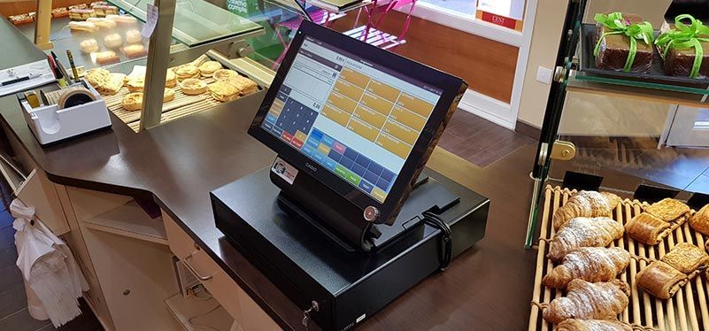 Un terminal de point de vente noir est posé sur le comptoir d'une boulangerie, à côté de plateaux de croissants et de viennoiseries fraîches.