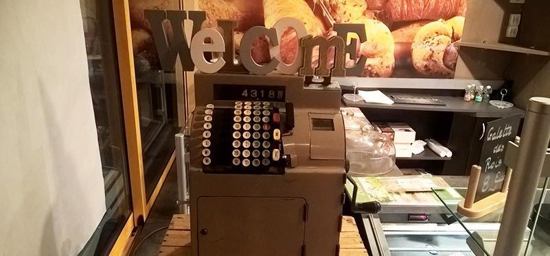 Dans une boutique ou un café, une caisse enregistreuse de style vintage est posée sur un comptoir devant un panneau « Bienvenue » fixé au mur.