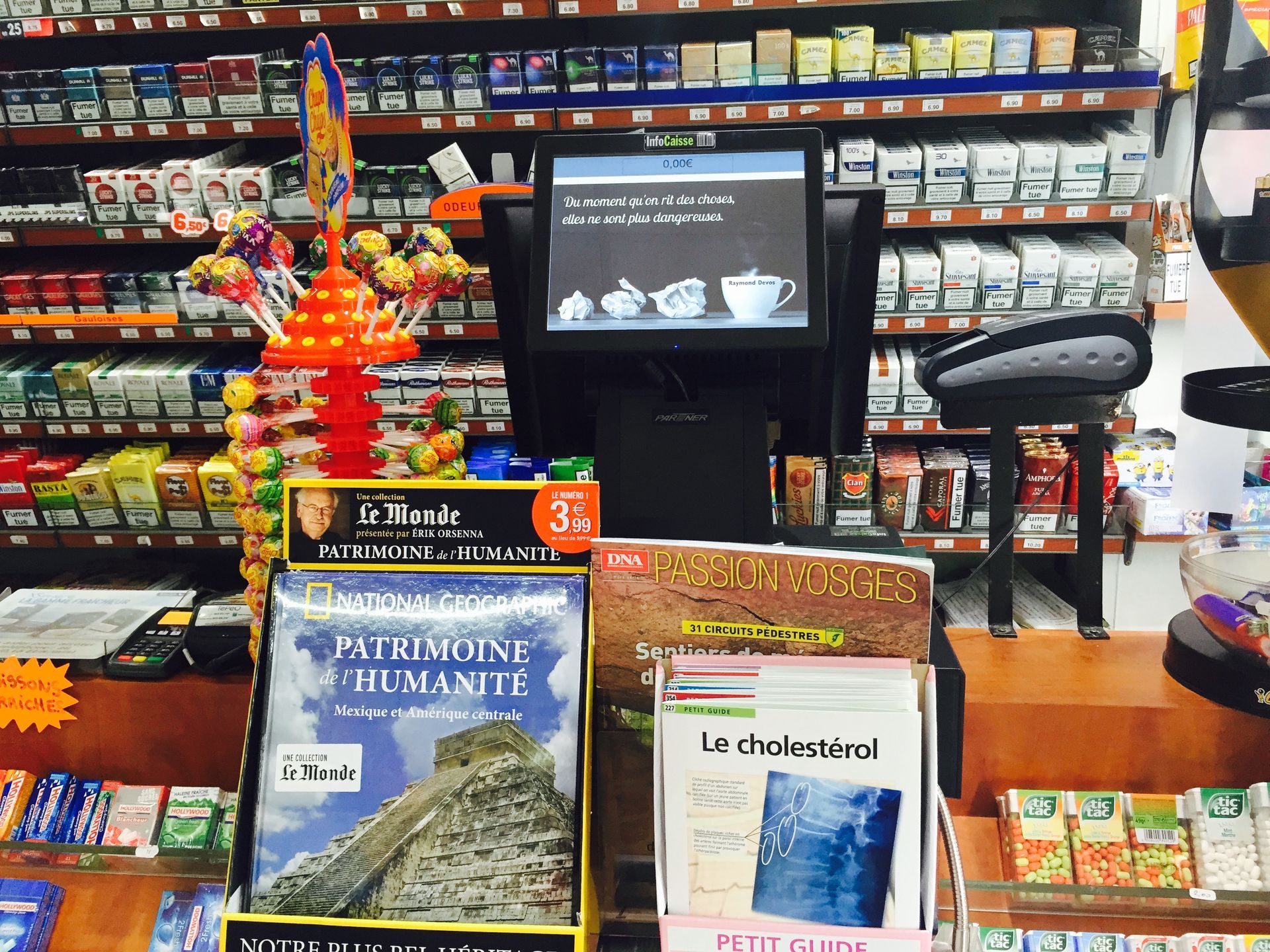 Le comptoir de caisse d'un magasin est équipé d'un écran d'ordinateur, de sucettes et de divers magazines, avec des étagères à cigarettes derrière.