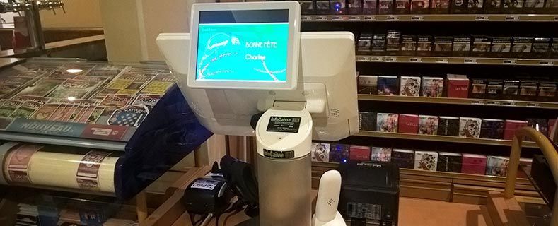 Un terminal de point de vente avec un écran vert est posé sur un comptoir dans un magasin dont on aperçoit des étagères remplies de produits en arrière-plan.