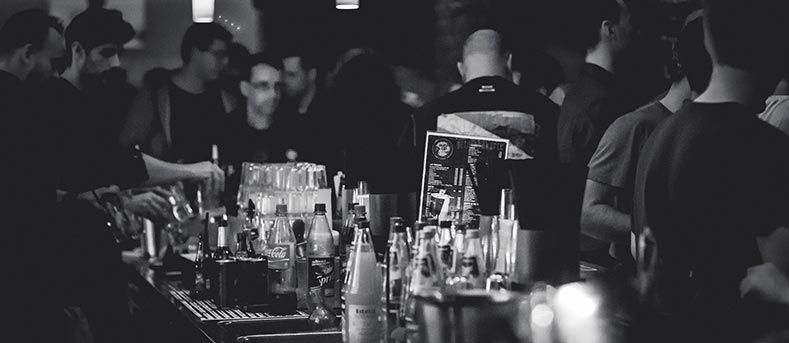 Vue en noir et blanc d'un bar bondé et faiblement éclairé, avec des gens debout autour d'un comptoir rempli de bouteilles de boissons.