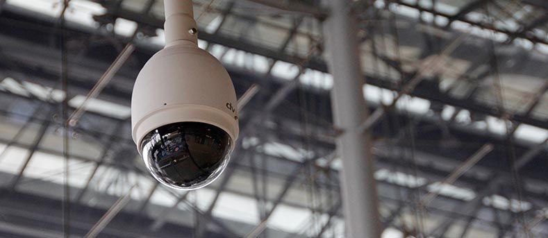 Une caméra de sécurité blanche en forme de dôme est suspendue à une structure de plafond sur un fond industriel lumineux.