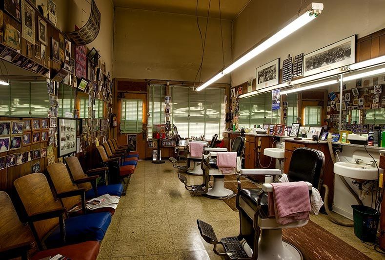 L'intérieur d'un salon de coiffure vintage, avec des rangées de chaises en bois, des fauteuils de barbier recouverts de nappes roses et des photos encadrées aux murs.