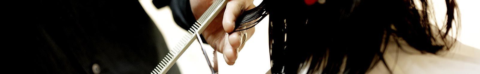 Une personne utilise des ciseaux en métal pour couper des cheveux foncés ; on aperçoit sa main et l'outil sur un fond blanc.