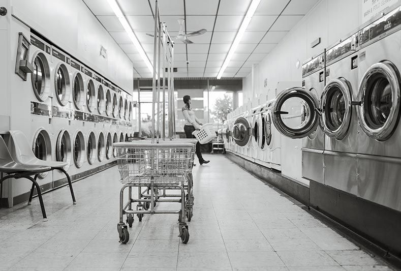 Une personne se tient debout dans une laverie automatique monochrome bordée de rangées de portes de machines à laver circulaires et de chariots à linge vides.