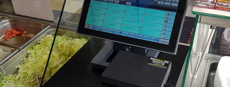 Un terminal de point de vente à écran tactile est posé sur un comptoir noir, à côté d'un récipient métallique pour la préparation des aliments rempli de laitue râpée.