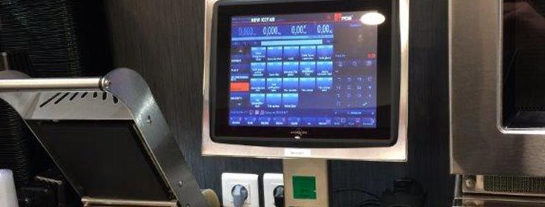 Un menu numérique tactile de point de vente, monté sur un support dans une cuisine ou un espace de restauration.