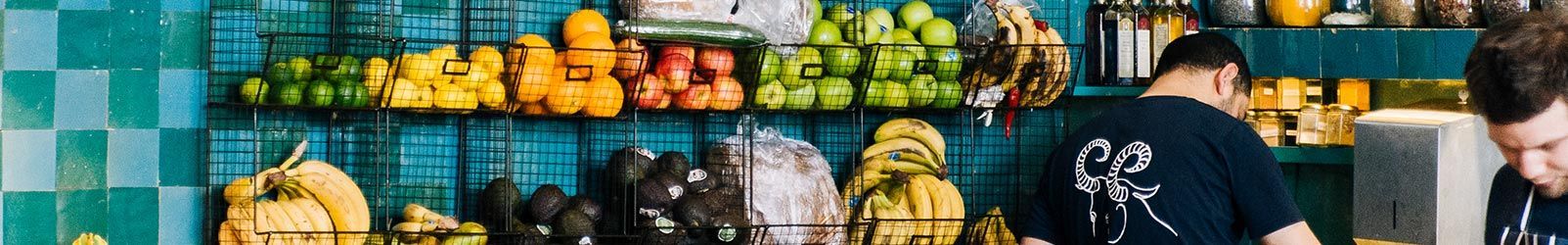 Un comptoir de cuisine avec des étagères de produits frais, notamment des agrumes, des pommes et des bananes, et deux ouvriers en arrière-plan.