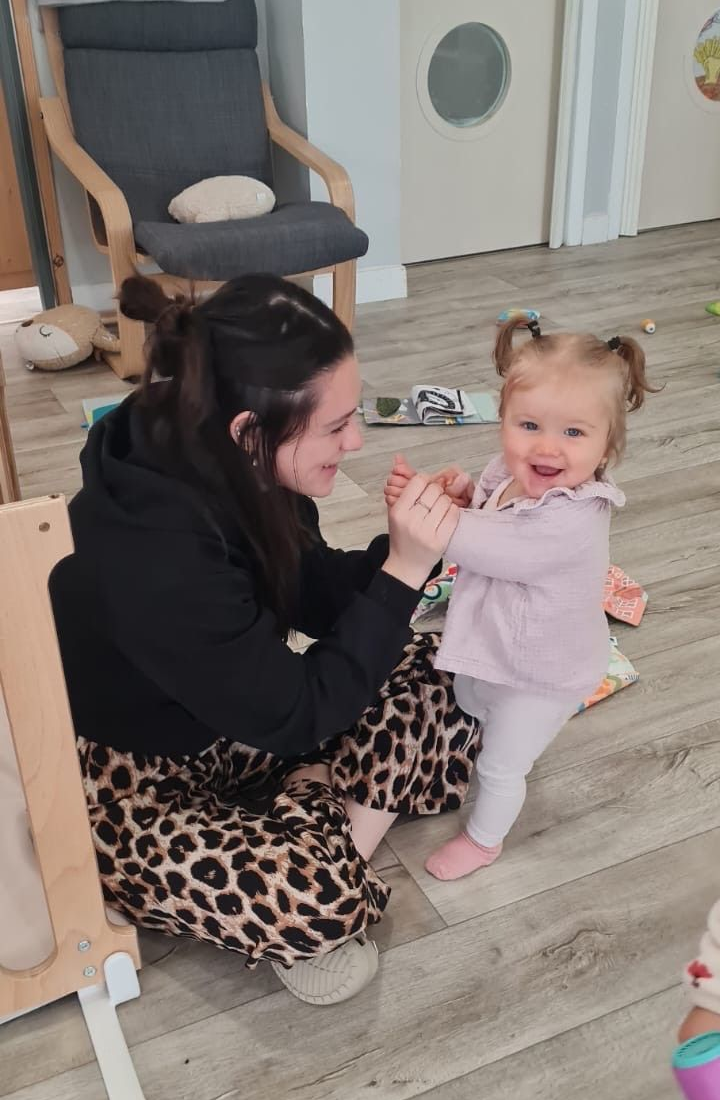 Une femme et un jeune enfant sourient en jouant. L'enfant est debout, la femme est agenouillée par terre.