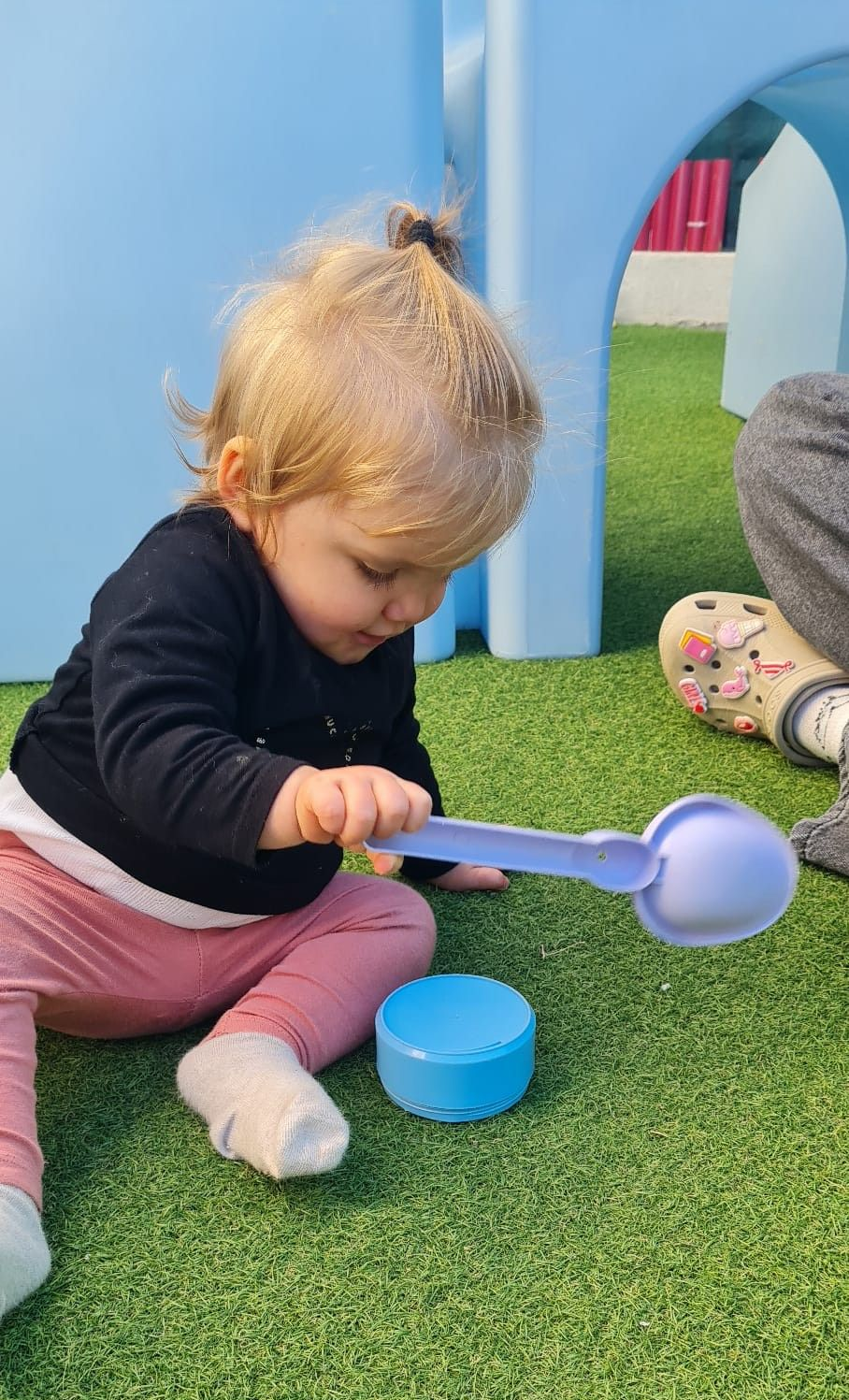 Enfant assis, jouant avec une cuillère bleue et un récipient sur une pelouse verte près d'une structure de jeu bleue.