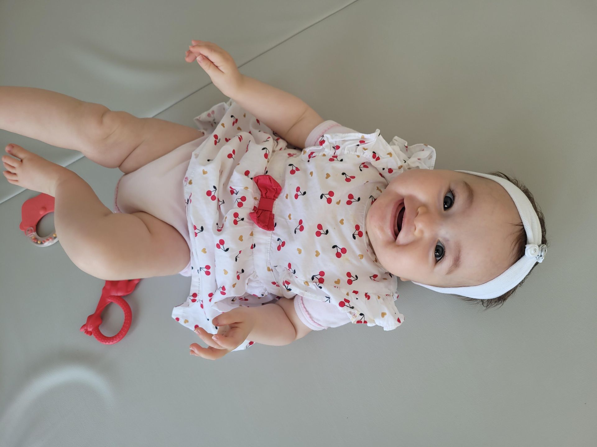 Bébé souriant, vêtu d'une tenue blanche à imprimés cerises avec un nœud rouge, allongé sur un tapis gris, tenant un jouet rouge.