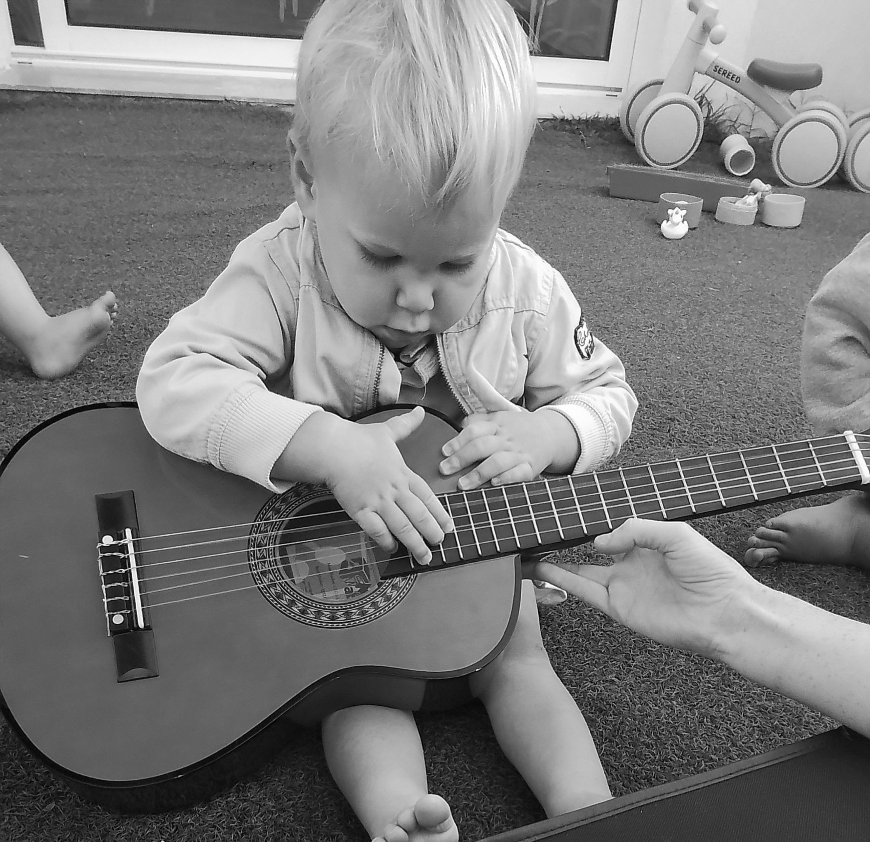Enfant assis, tenant et explorant une petite guitare, guidé par la main d'un adulte.