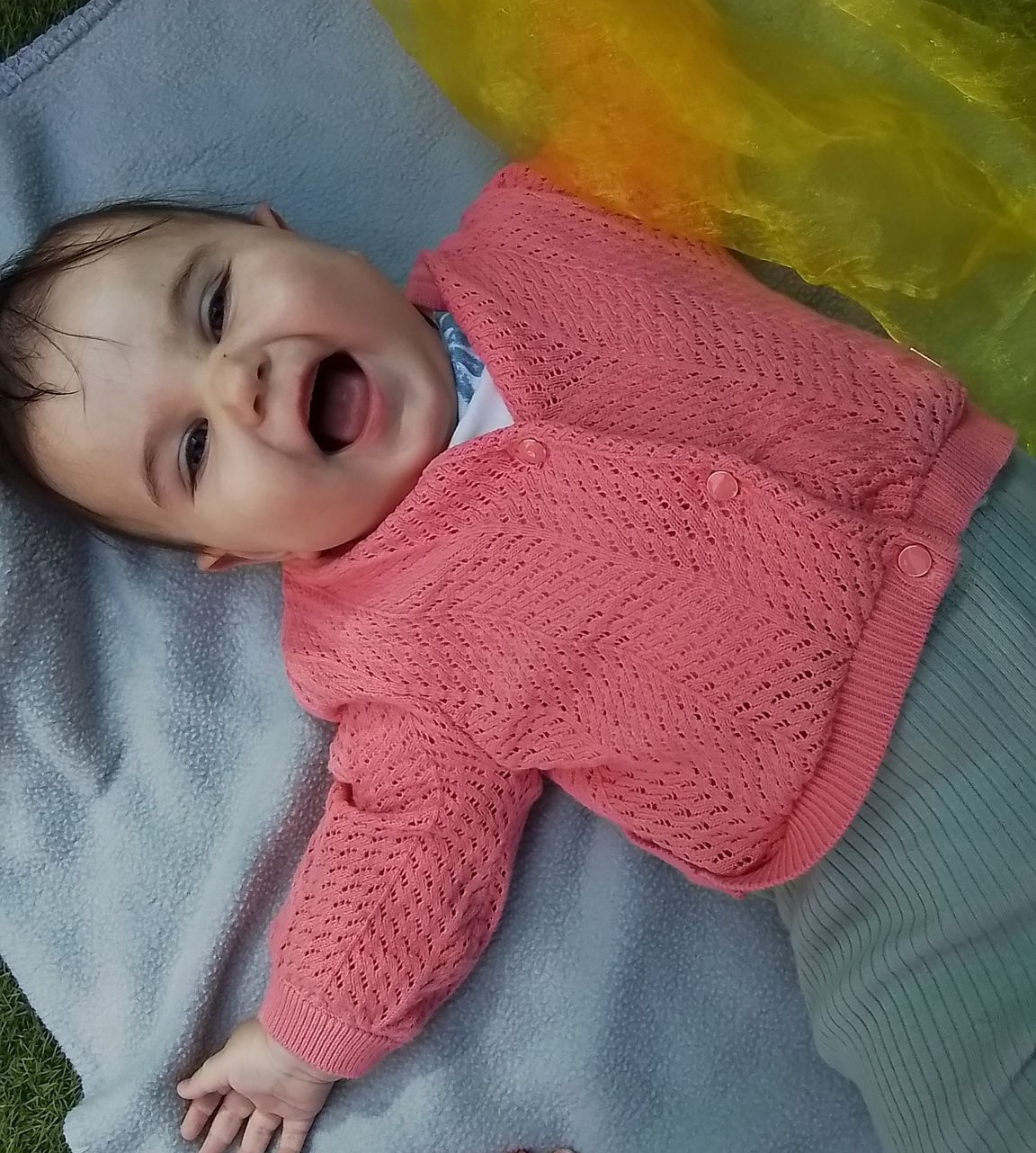 Bébé portant un pull rose en tricot, souriant et riant dehors sur une couverture.