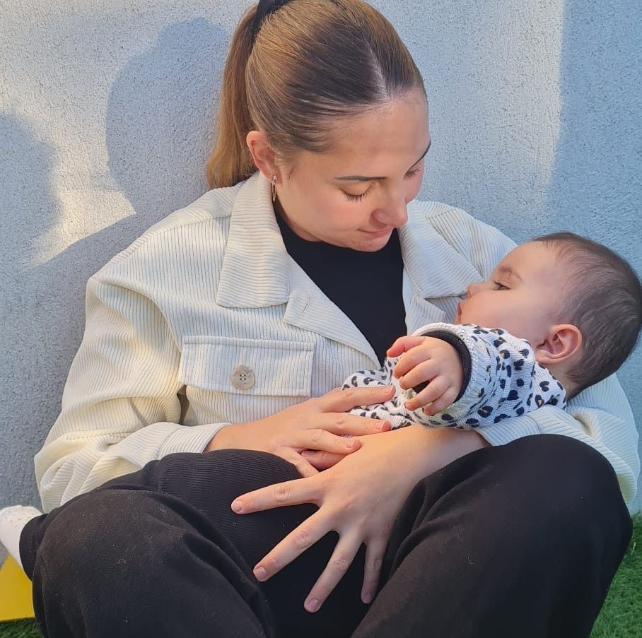 Femme aux cheveux attachés en queue-de-cheval tenant un bébé dans ses bras, le regardant avec tendresse. Bébé vêtu d'une tenue à motifs. Scène en extérieur.