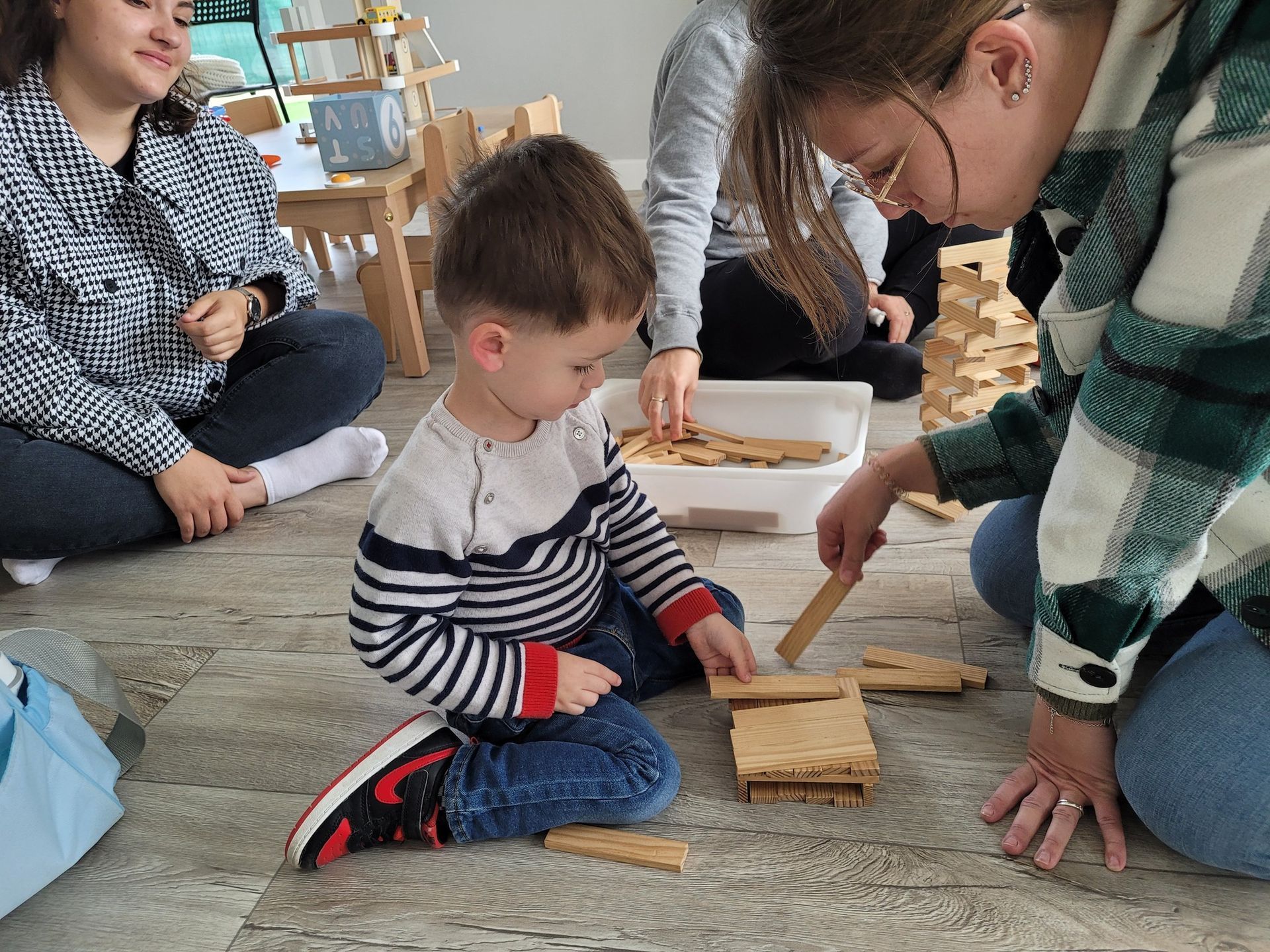 Un enfant et un adulte jouent avec des blocs de bois sur le sol, d'autres personnes sont visibles en arrière-plan.