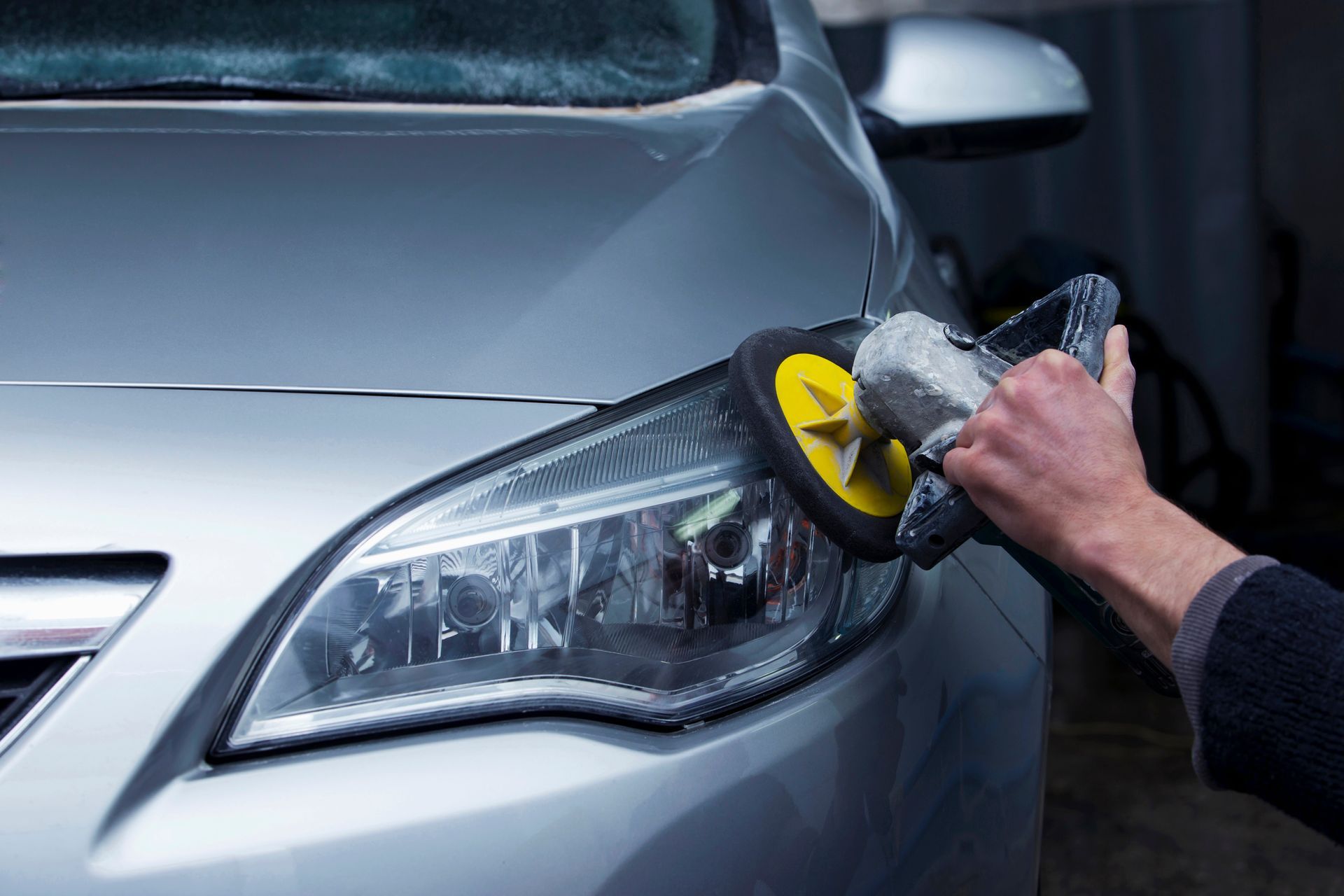 Personne nettoyant un phare de voiture argenté avec un outil de nettoyage jaune et gris.