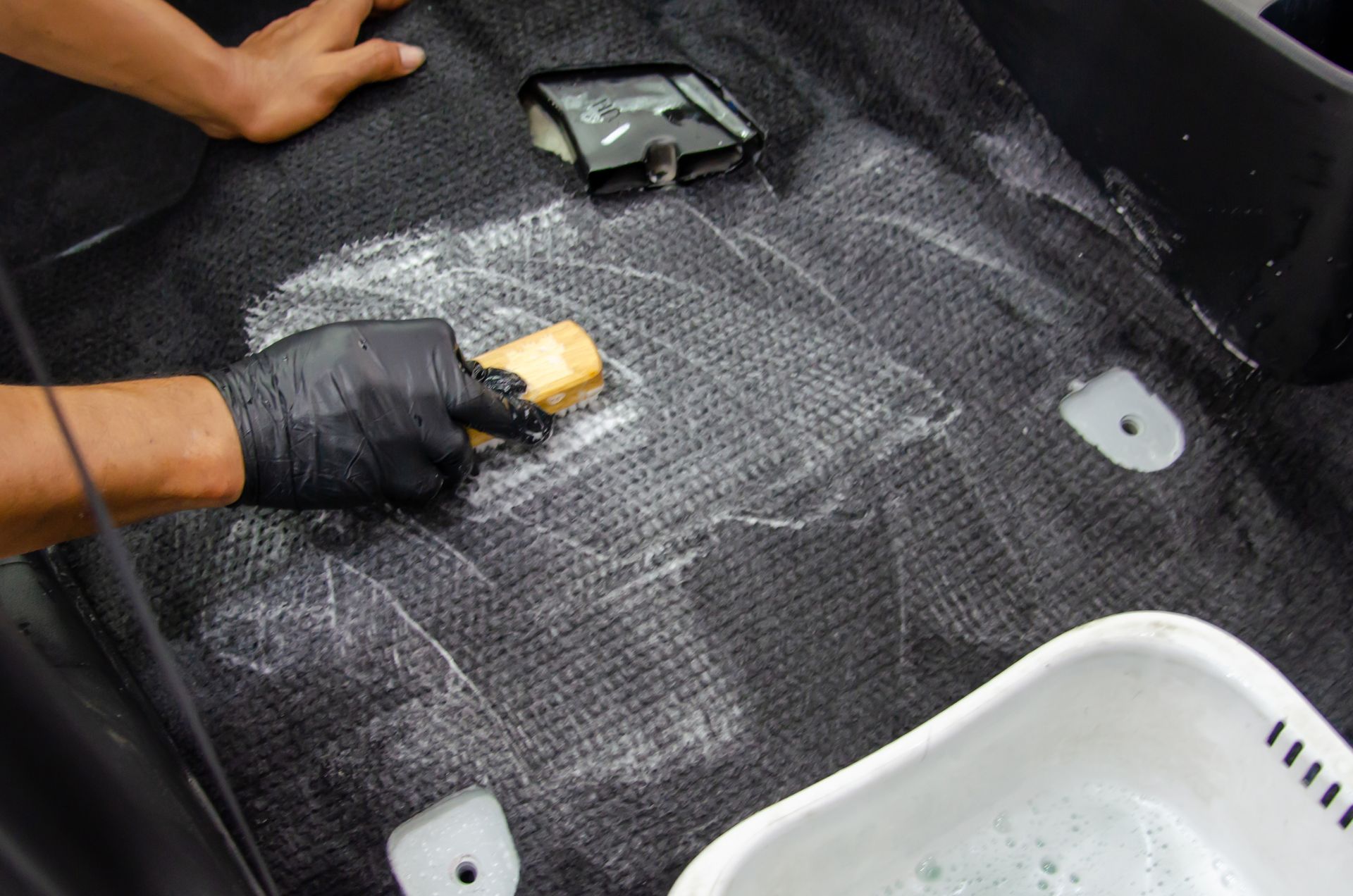 Un homme portant un gant noir frotte le sol de la voiture avec une brosse. De la mousse blanche recouvre le tapis sombre.