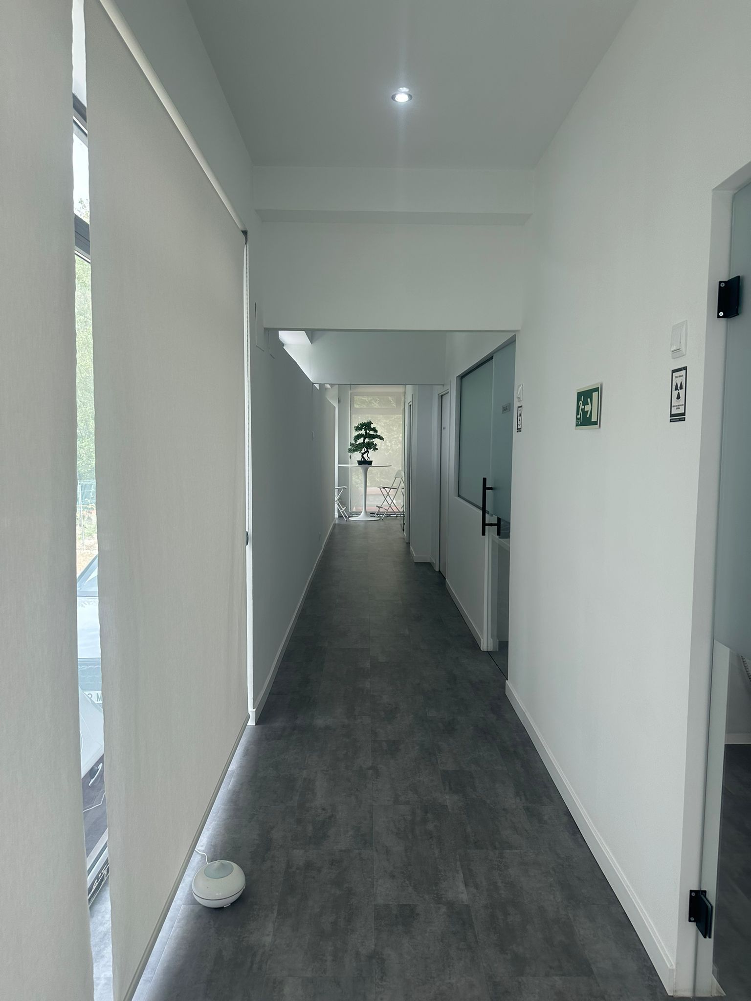 Pasillo estrecho de color blanco con suelo gris, puertas a la derecha y una ventana al final.