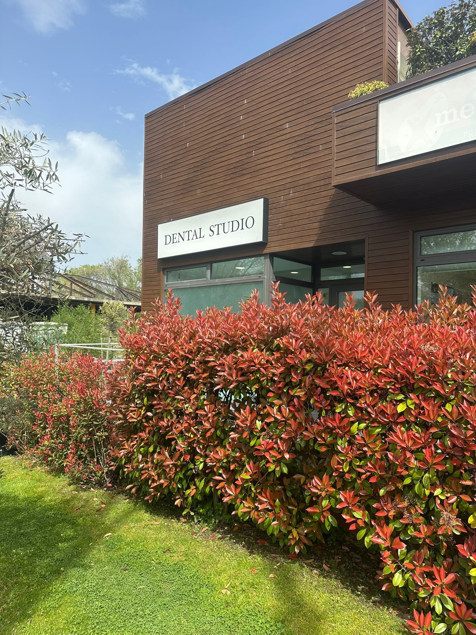 Edificio de ladrillo con letrero blanco y entrada de cristal, enmarcado por setos rojos y césped verde bajo un cielo parcialmente nublado.