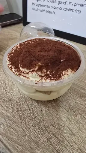 Tiramisú servido en un bol transparente, espolvoreado con cacao en polvo, sobre la mesa de una cafetería.