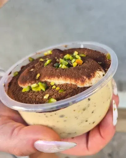 Mano sosteniendo una taza transparente de postre tiramisú cubierto con cacao en polvo y pistachos picados.