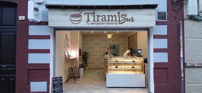 Fachada de una tienda de tiramisú con entrada abierta, vitrina iluminada y asientos en el interior.