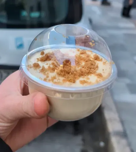 Una mano sostiene un vasito de postre de plástico transparente cubierto con crema blanca y migas al aire libre.