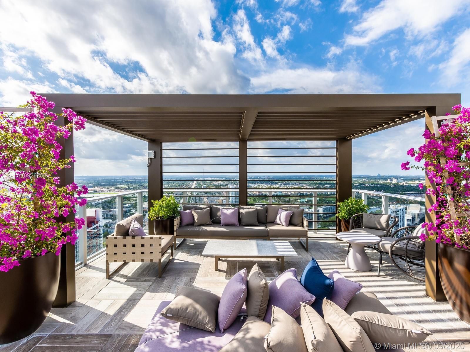 A rooftop deck with a couch , table , chairs and flowers.