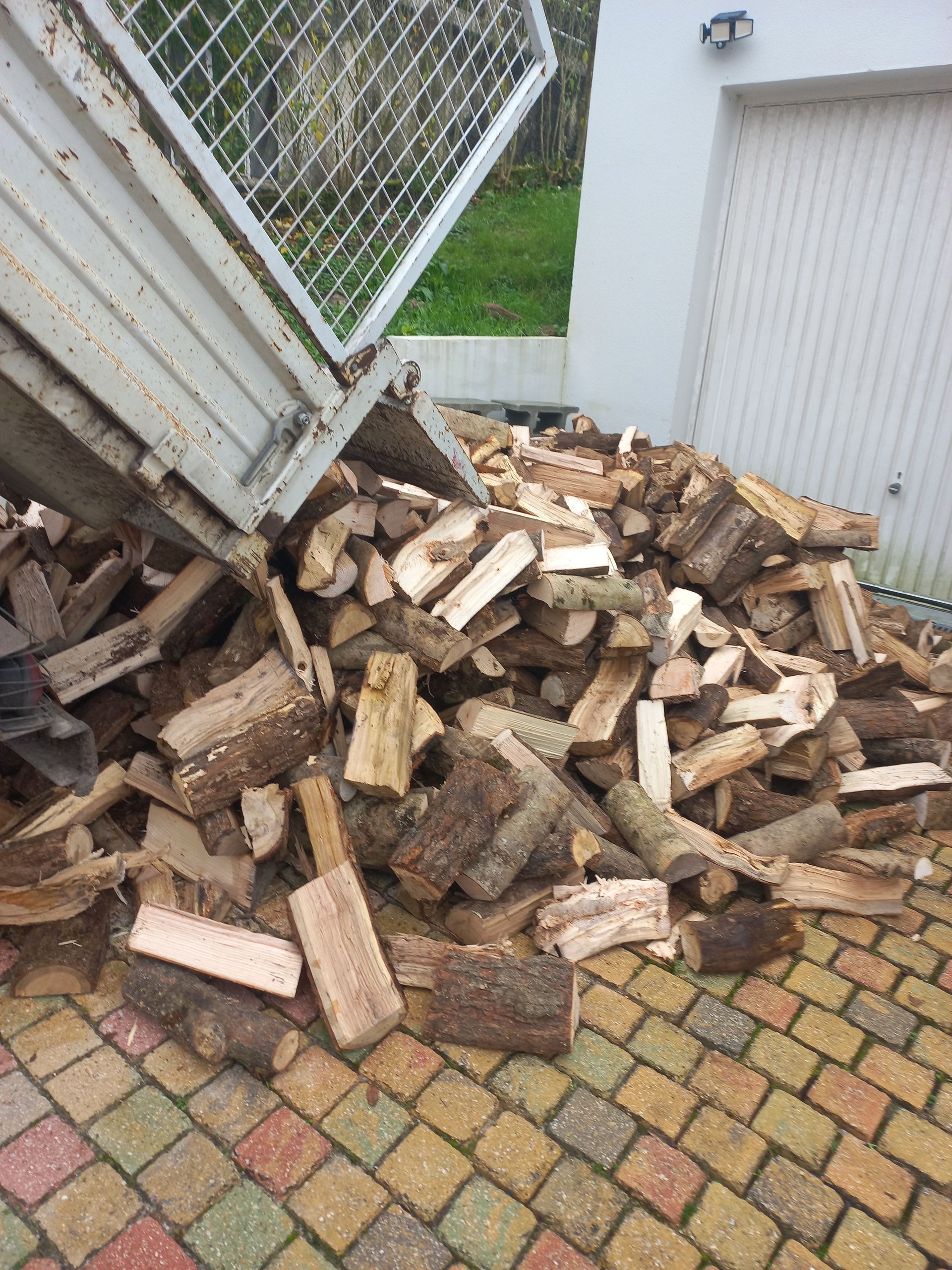 Un camion-benne décharge une grande pile de bois de chauffage coupé sur une allée pavée à côté d'un bâtiment blanc.