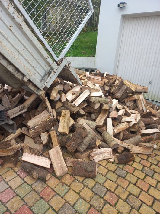 Un camion-benne décharge une grande pile de bois de chauffage coupé sur une allée pavée à côté d'un bâtiment blanc.