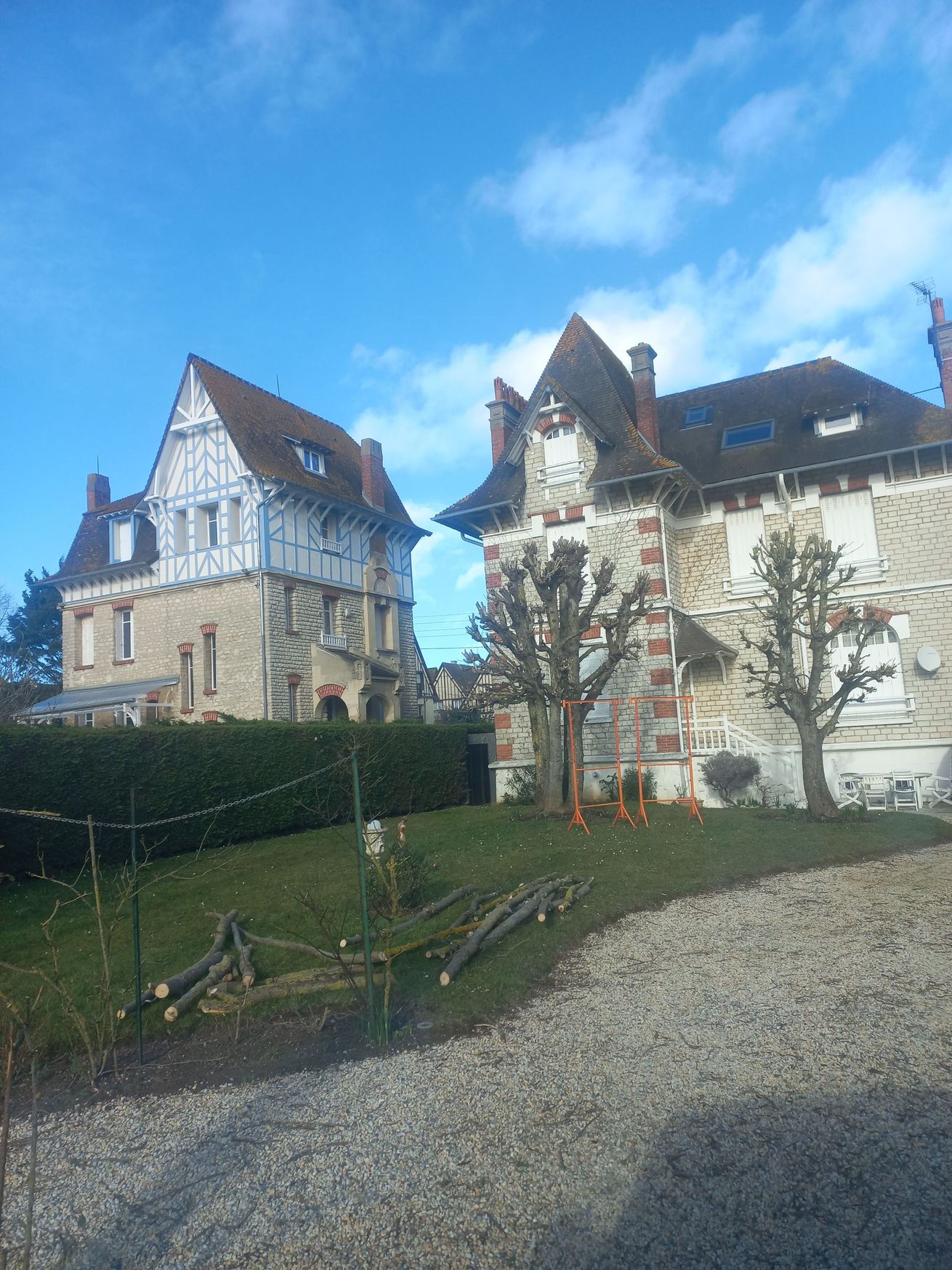 Deux maisons traditionnelles normandes en pierre aux toits de tuiles se dressent par une journée ensoleillée derrière une pelouse verte et une allée de gravier.