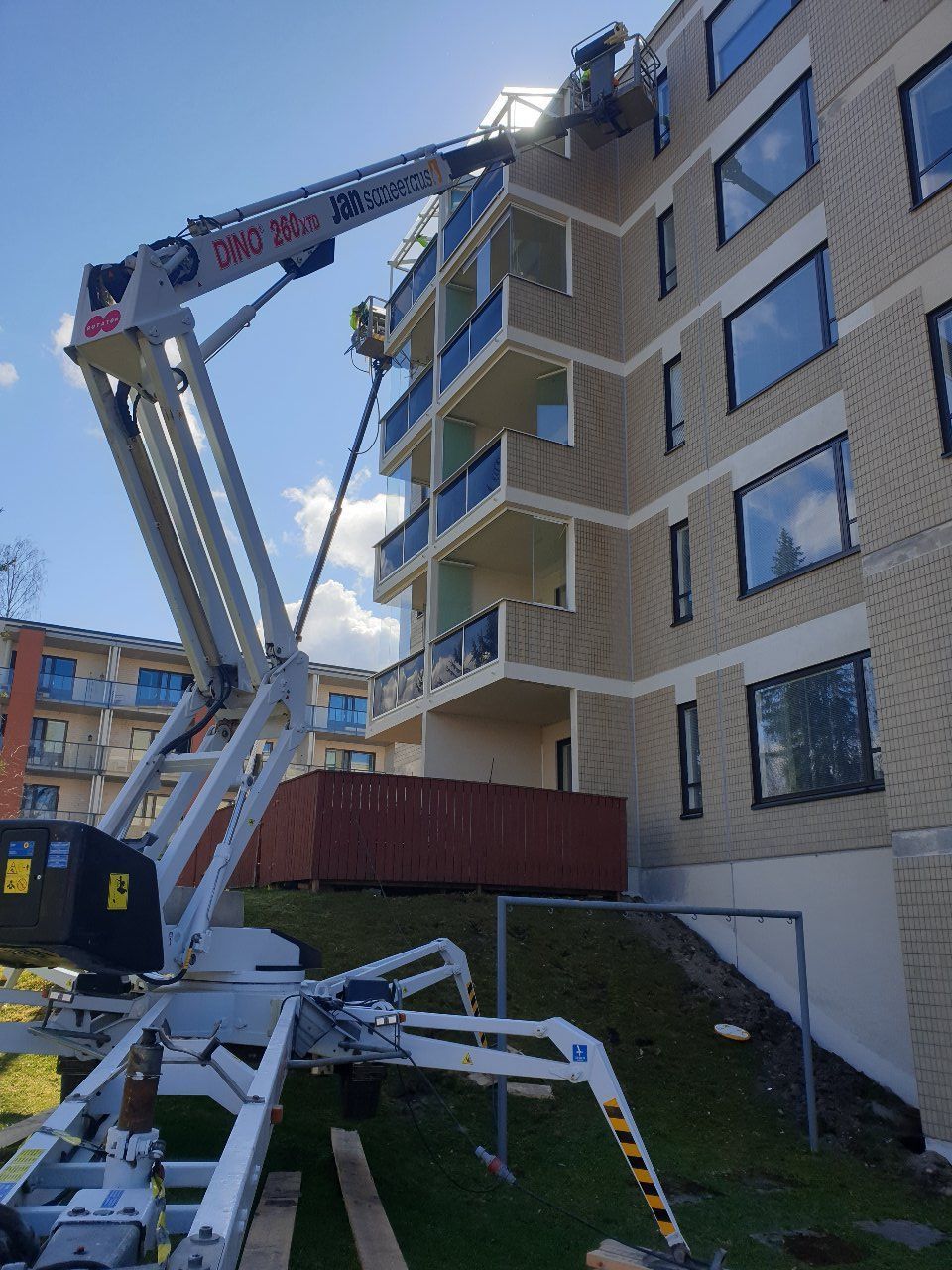 Nosturi nostaa materiaaleja beigenvärisen tiilikerrostalon seinustalle aurinkoisena päivänä.