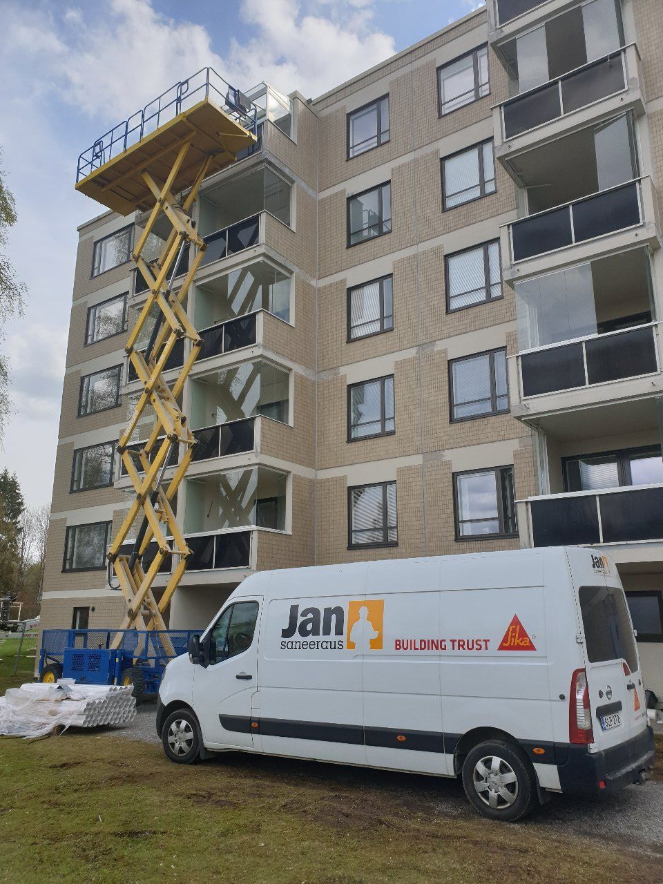 Valkoinen pakettiauto logolla pysäköitynä monikerroksisen rakennuksen viereen. Saksinostin parvekkeiden lähellä.