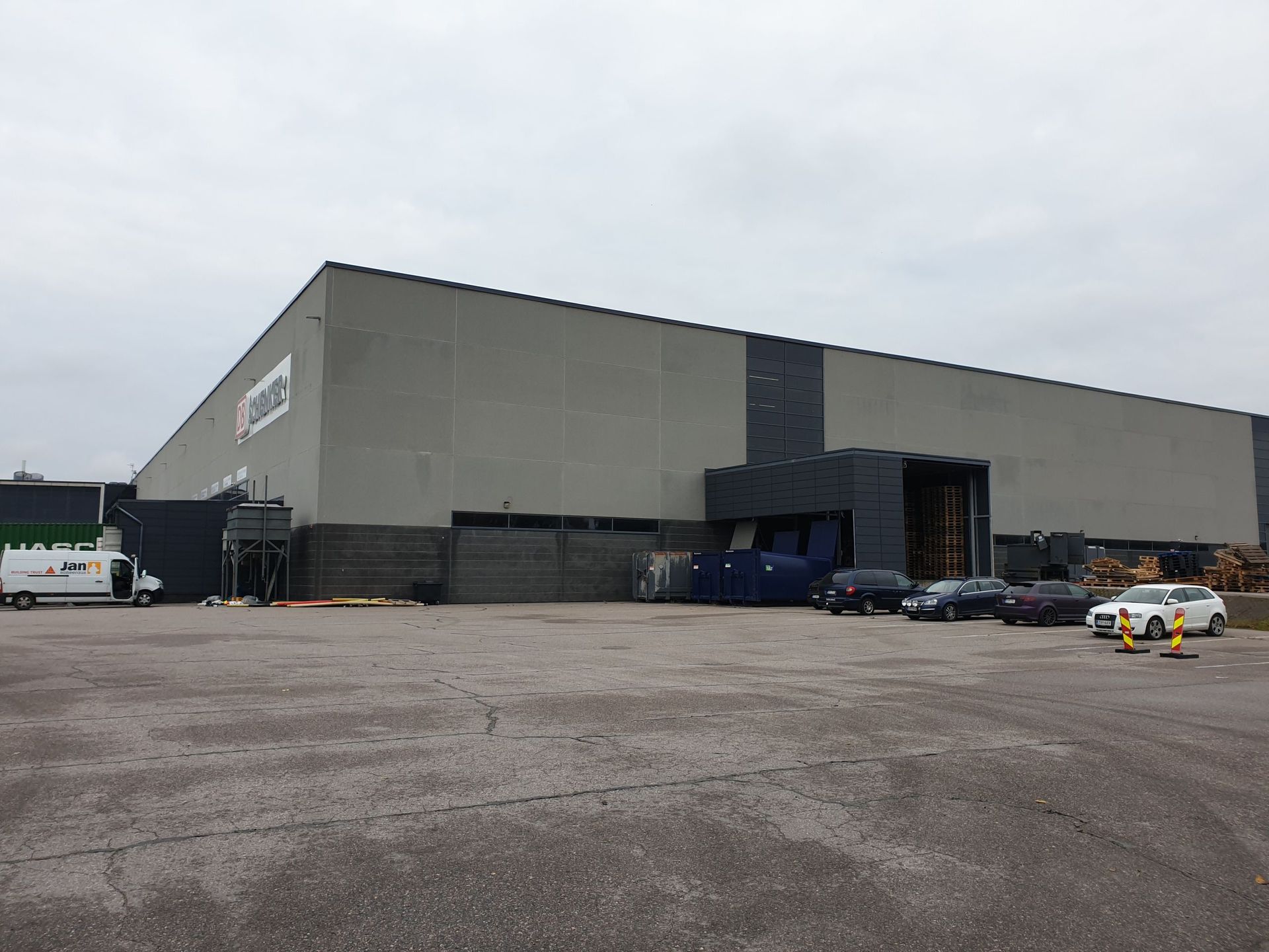 Suuri harmaa teollisuusrakennus, jossa on pysäköityjä ajoneuvoja ja kuorma-auto harmaana päivänä.