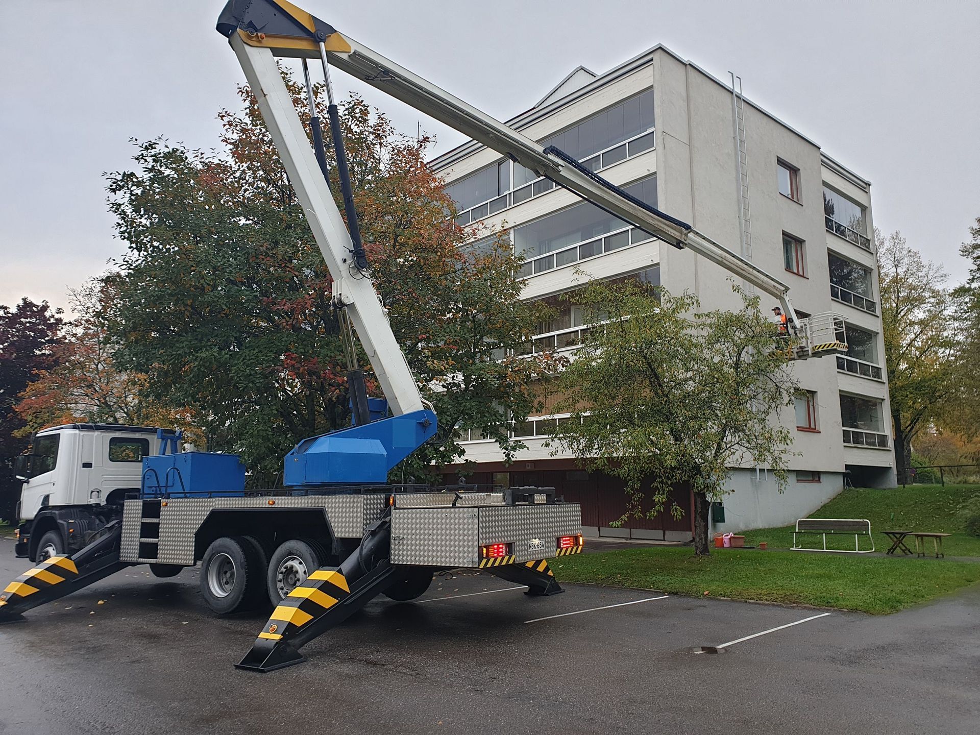 Nosturiauto nostamassa kuormaa monikerroksisen rakennuksen lähellä. Sininen nosturin varsi ulottuu kohti parveketta. Valkoinen kuorma-auto, harmaa rakennus.
