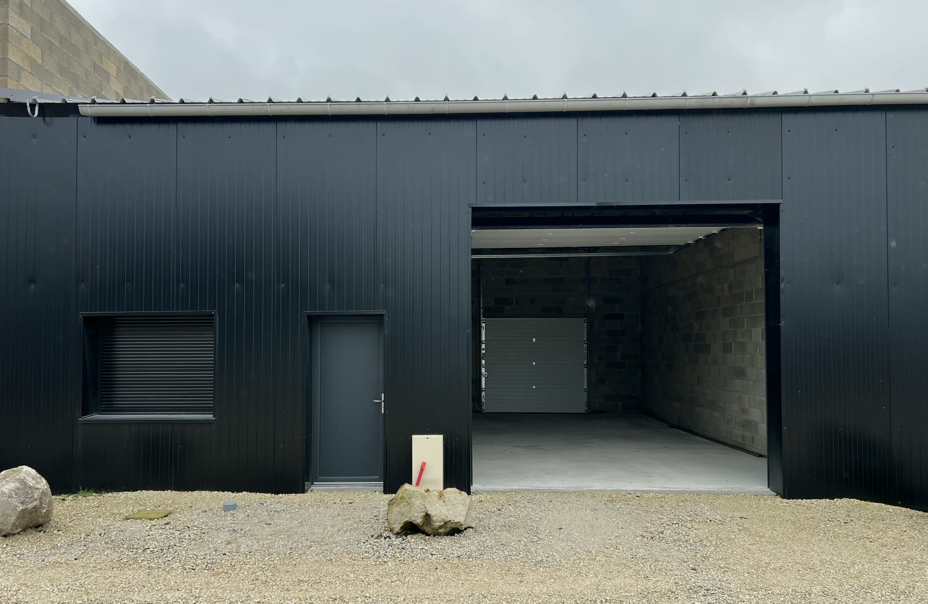 Garage noir avec entrée ouverte, petite porte et aération. Allée en gravier.