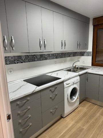 Una cocina con gabinetes grises, encimeras blancas, lavadora y secadora, y un fregadero.