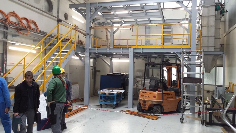 Intérieur d'une installation avec un chariot élévateur, des ouvriers, une plate-forme surélevée et des machines.