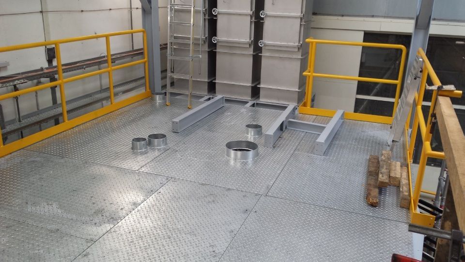 Plateforme métallique avec garde-corps jaunes, structures métalliques verticales et objets cylindriques au sol.