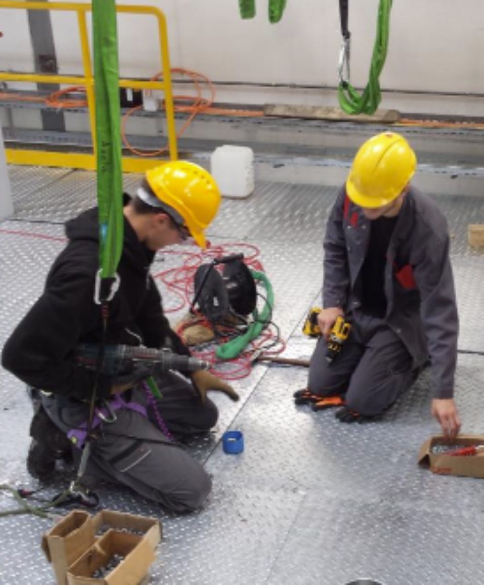 Deux ouvriers portant des casques de chantier percent sur un sol métallique. L'un d'eux est suspendu.