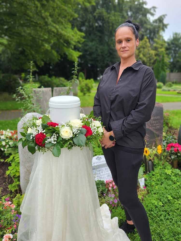 Eine Frau steht auf einem Friedhof vor einer mit Blumen geschmückten Urne.