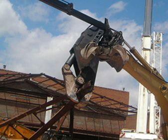 Un brazo de excavadora hidráulica amarillo con un accesorio de cizalla para demolición de metal, recortado contra un cielo azul nublado.