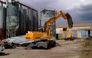 Una excavadora de construcción amarilla con una garra incorporada, estacionada en un terreno industrial con grandes silos metálicos.
