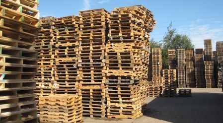 Grandes pilas de palés de madera desgastados por el tiempo se alinean en un patio al aire libre bajo un cielo azul despejado.