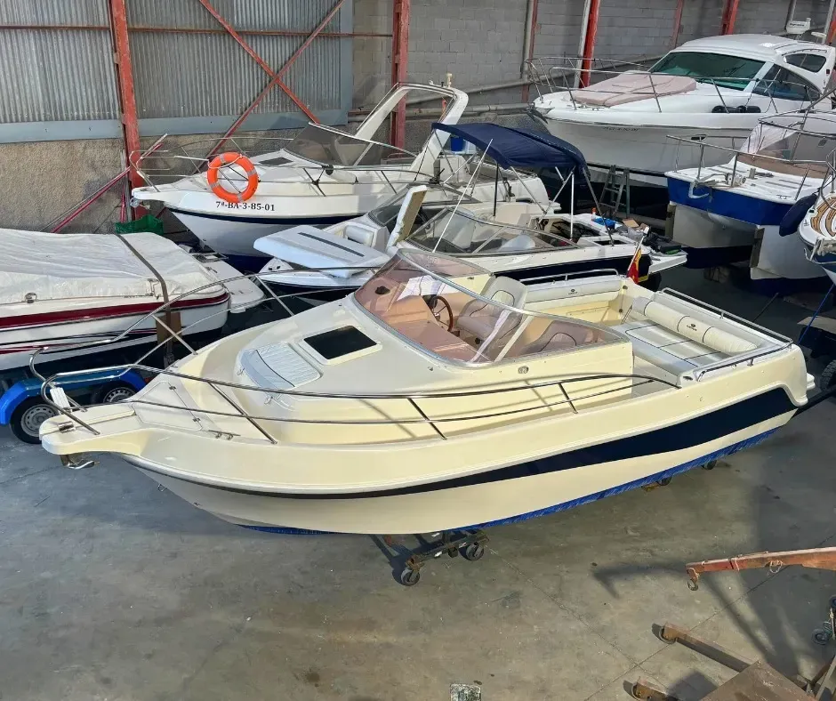 Una lancha motora de color beige con una franja azul se encuentra en un almacén, rodeada de otras embarcaciones y equipos.