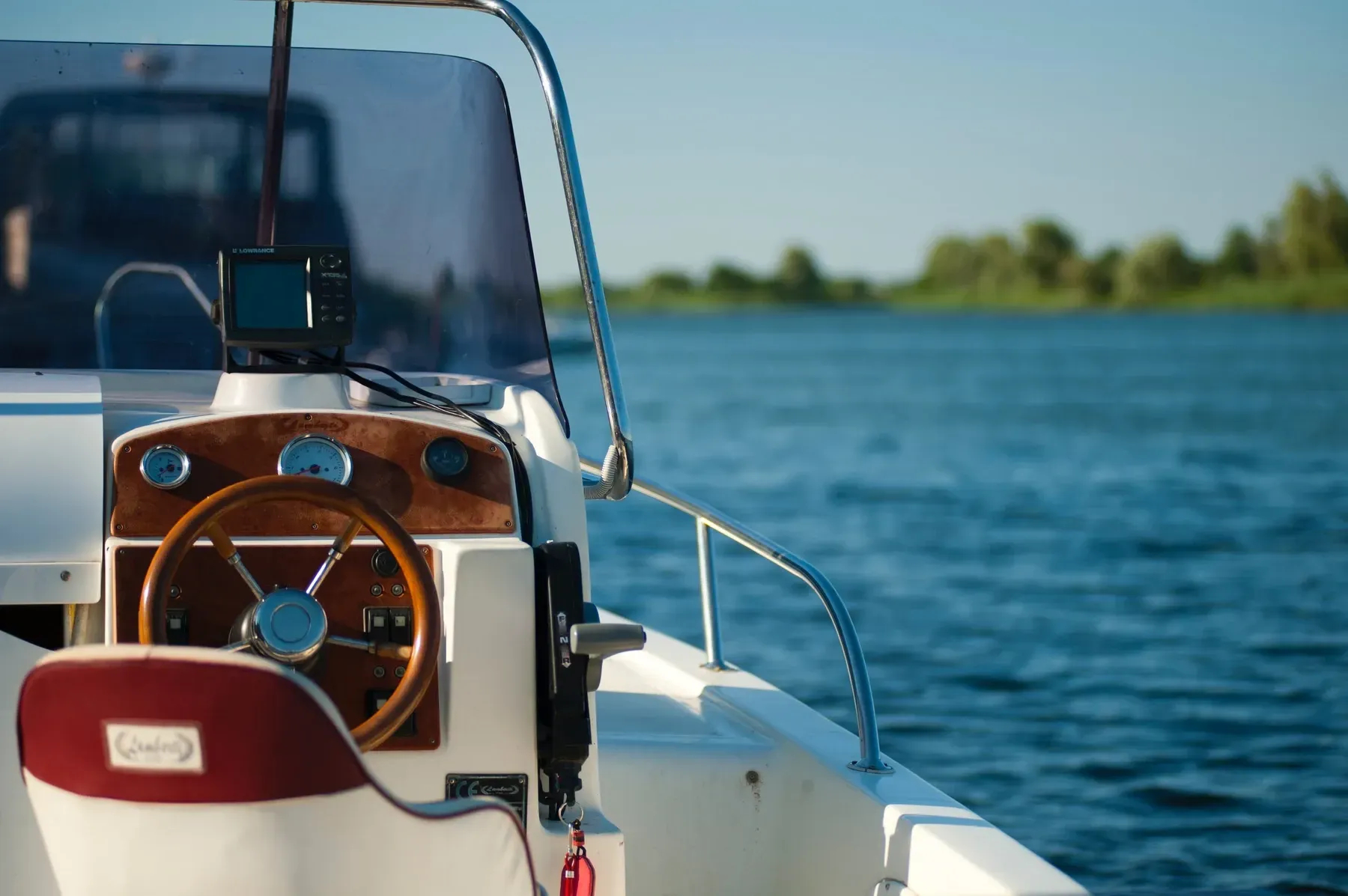 Timón de barco con volante de madera, vista al agua y a la orilla lejana.