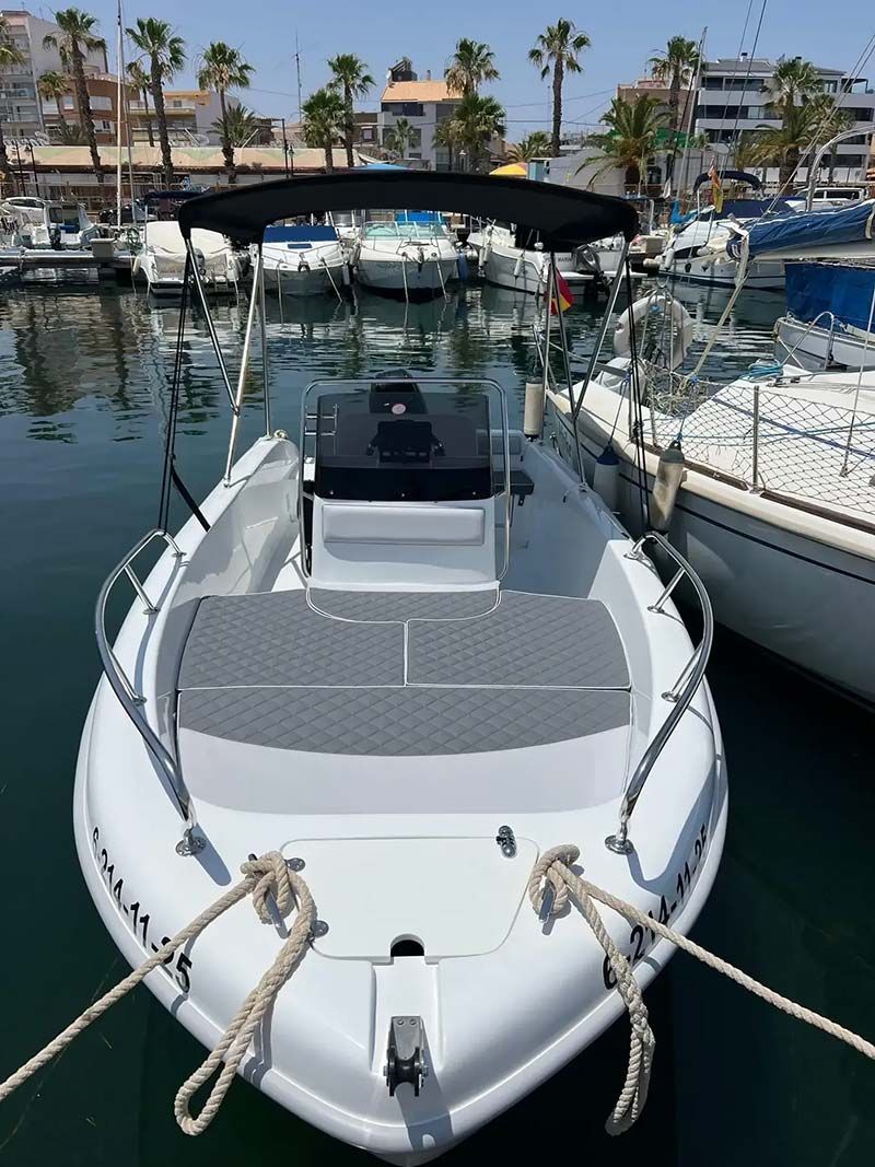 Lancha motora blanca atracada en un puerto deportivo, vista desde la proa con edificios ribereños bordeados de palmeras al fondo.